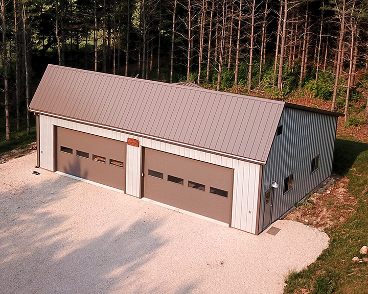 Post-frame detached garage with steel siding, two overhead doors, and gravel driveway in wooded setting