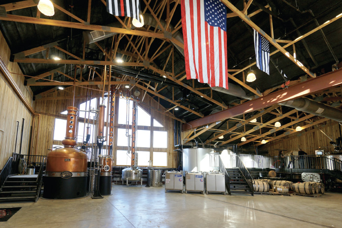 Interior of a post frame distillery with exposed wood trusses, copper stills, American flags, large windows, and open production floor. American Flag Hanging