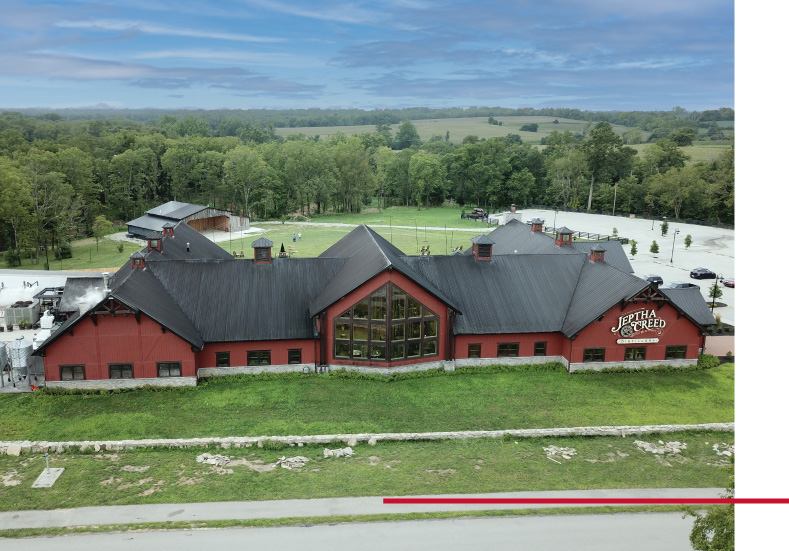 Aerial view of a red post frame distillery building with black metal roof, large central windows, and Jeptha Creed Distillery signage.