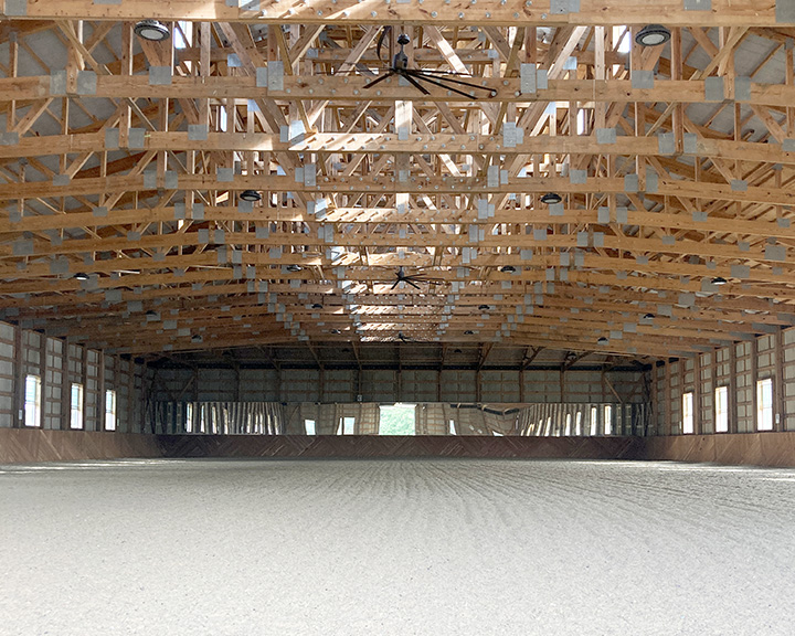 Clear-span indoor riding arena interior with wood trusses, steel walls, and riding surface footing