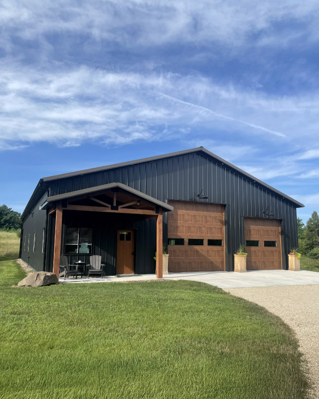 Post frame shop building with steel siding, timber frame porch, overhead garage doors, and windows