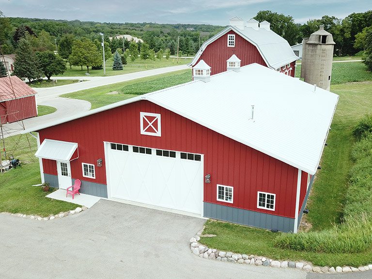 Post-frame hobby workshop with red steel siding, overhead garage door, cupolas, and adjacent barn in rural setting
