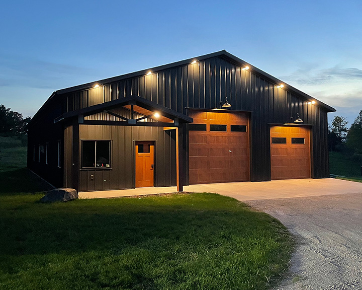 Post-frame hobby workshop garage with black steel siding, overhead doors, covered entry, and exterior lighting at dusk