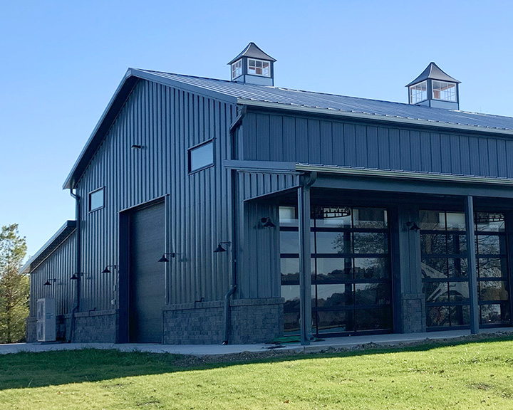 Post-frame hobby workshop with steel siding, glass overhead doors, covered porch, and cupolas