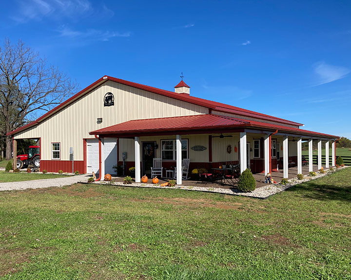 Farmhouse style barndominium with steel siding, red steel roof, wraparound covered porch, and rural setting