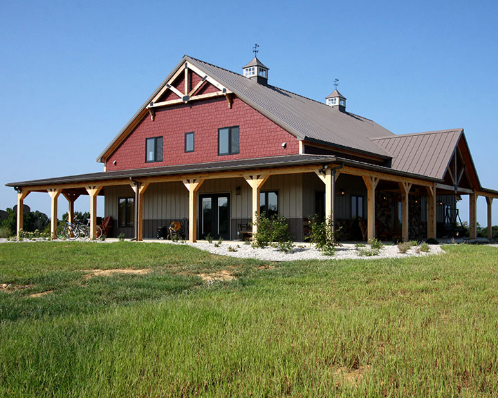 Farmhouse barndominium with steel siding, wraparound porch, timber posts, and cupolas on rural property