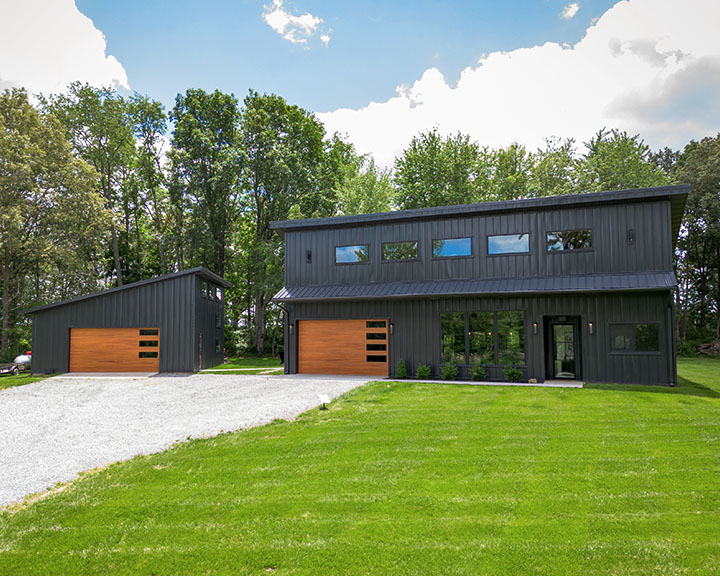 Modern barndominium with black steel siding, large windows, and detached garage with wood overhead doors on rural property