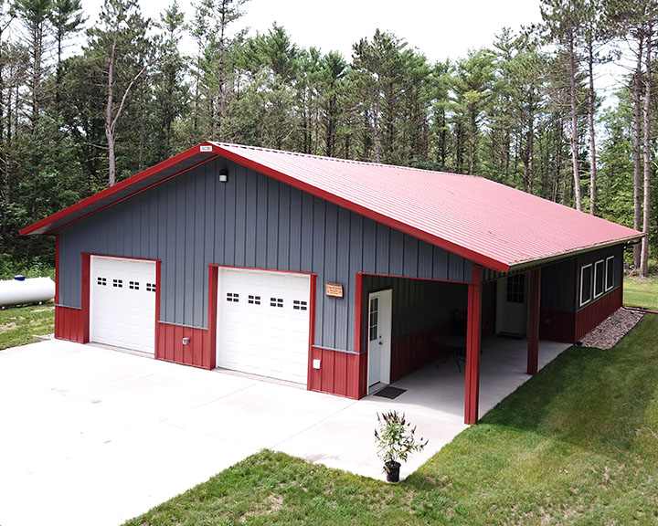 Post-frame cabin garage with red steel roof, gray steel siding, overhead doors, and covered porch in wooded setting