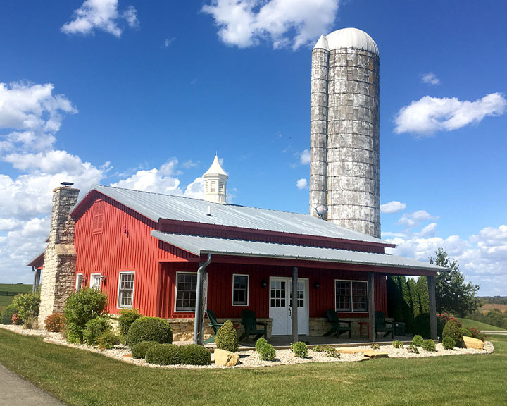 Post-frame cabin hunting lodge with red steel siding, cupola, covered porch, and adjacent silo in rural setting