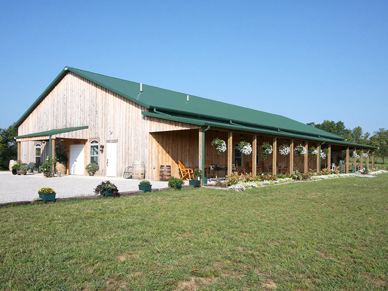 Custom Event Barn with Covered Porch and Open-Span Interior Post frame event barn with wood siding, green steel roof, and covered porch designed for wedding venues and community events