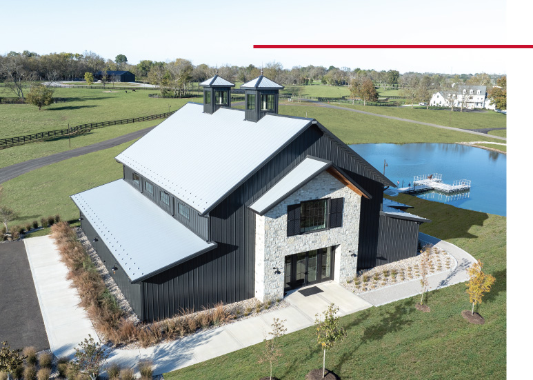 Keene Ranch Event Barn Aerial View | Custom Post Frame Event Barn Aerial view of the Keene Ranch event barn with black steel siding, stone entry facade, cupolas, and pond backdrop.