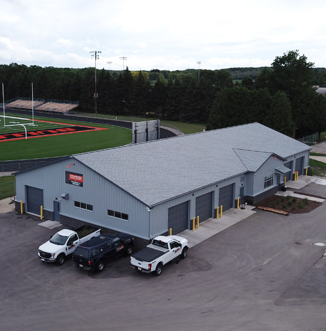 Maintenance garage with steel siding, multiple overhead doors, and attached office near sports field