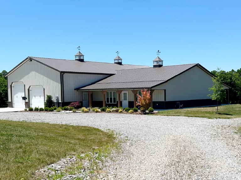 Post frame barndominium with attached garage, steel siding, multiple cupolas, and residential entry on rural property