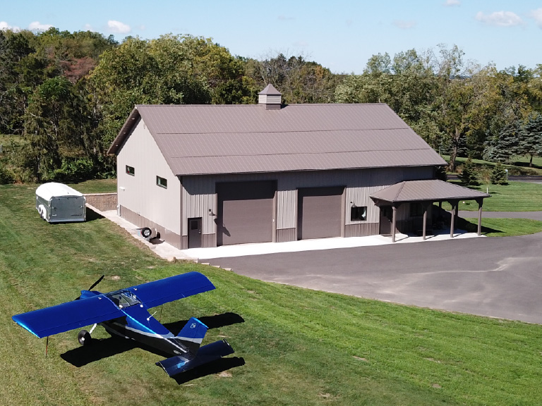 Post frame airplane hangar with steel siding, large overhead doors, and covered porch on rural property with small aircraft