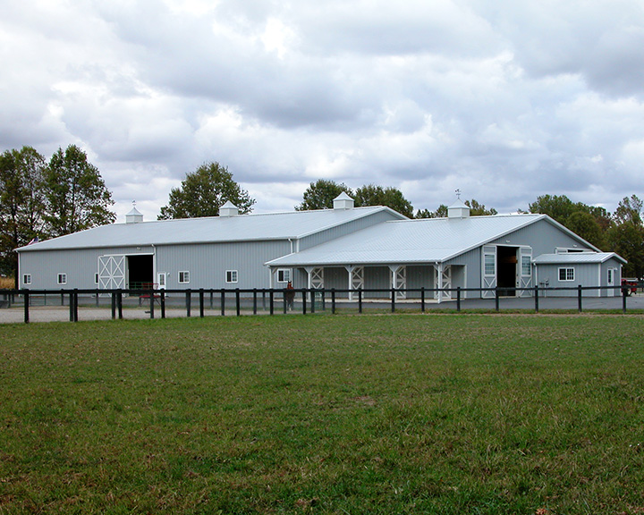 Indoor horse riding arena with attached stable, steel siding, cupolas, and covered porch