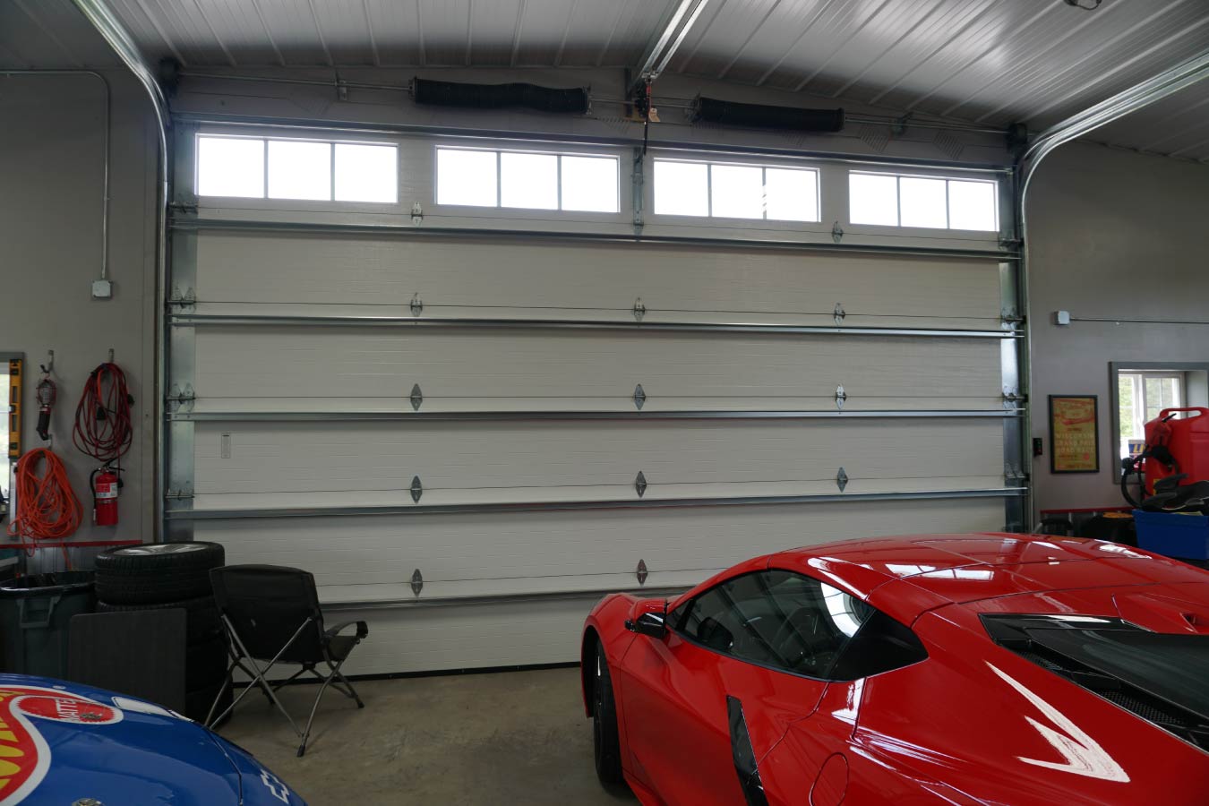 This car collection garage showcases a refined blend of traditional barn aesthetics and modern functionality through post frame construction. The red steel siding paired with white trim and a gray wainscot creates a clean, durable exterior designed to stand the test of time. Along the upper portion of the sidewall, a row of clerestory-style windows brings in natural light while maintaining privacy—ideal for illuminating a vehicle collection without sacrificing wall space for storage or displays. The white metal roof enhances energy efficiency and longevity, while the cupola adds both ventilation and architectural character. The structure also features a large overhead door and a separate entry door with a protective awning, offering convenient access for both vehicles and everyday use. Surrounding site elements, including the curved driveway and landscaping, highlight how a car collection garage can be seamlessly integrated into a property’s layout. Built with post frame construction, this garage provides wide-open interior space, making it perfect for storing, showcasing, and working on vehicles with maximum flexibility and efficiency.