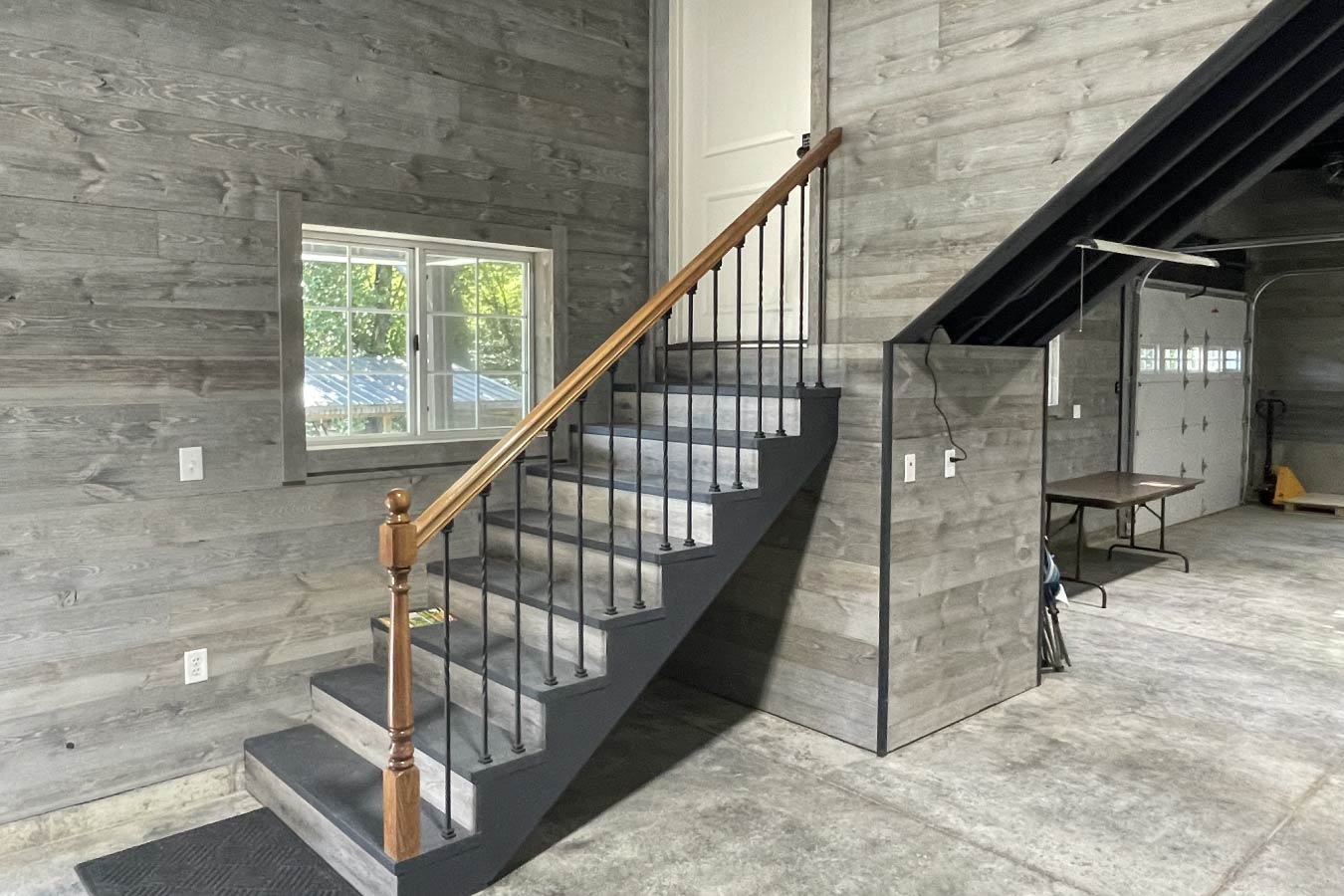 This staircase inside a garage with living quarters provides convenient access between the garage level and the upper living space. Built within a post frame structure, the layout allows for efficient use of space while maintaining a clean and functional design. As with all Walters Buildings projects, both the siding and roof are constructed from steel (unless otherwise specified), ensuring long-term durability on the exterior while allowing for fully finished interior spaces. The post frame construction enables flexible layouts, making it easy to integrate features like this interior stairway without compromising open space below. The staircase features carpeted steps, a wood handrail, and a well-lit landing, enhancing both safety and comfort. Its placement allows for seamless movement between levels, supporting the dual-purpose design of the garage with living quarters. This detail highlights how post frame construction can effectively support both utility and residential functionality within a single building.