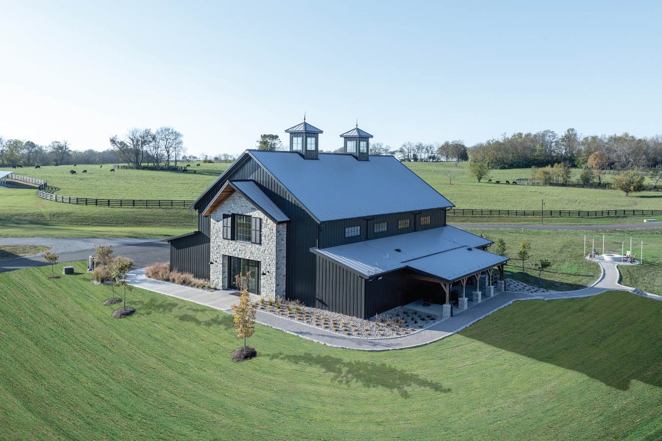 41-3955 Keene Ranch event barn with black steel siding, stone entry facade, covered porch, and dual cupolas overlooking rolling pasture.
