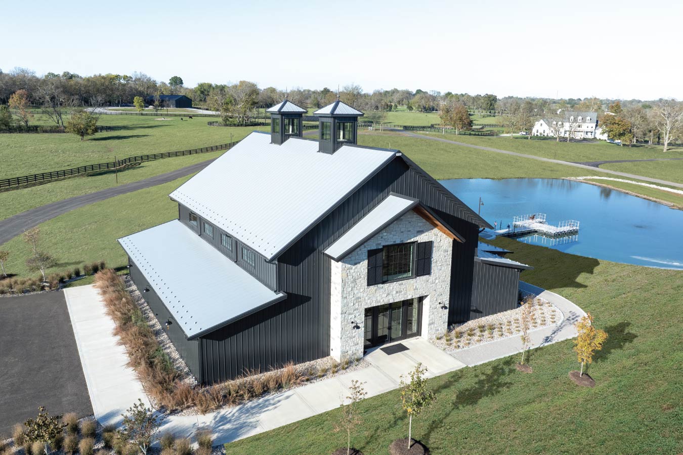 Keene Ranch event barn with black steel siding, stone entry facade, metal roof, and dual cupolas overlooking a pond. Pond with Path from Event Barn
