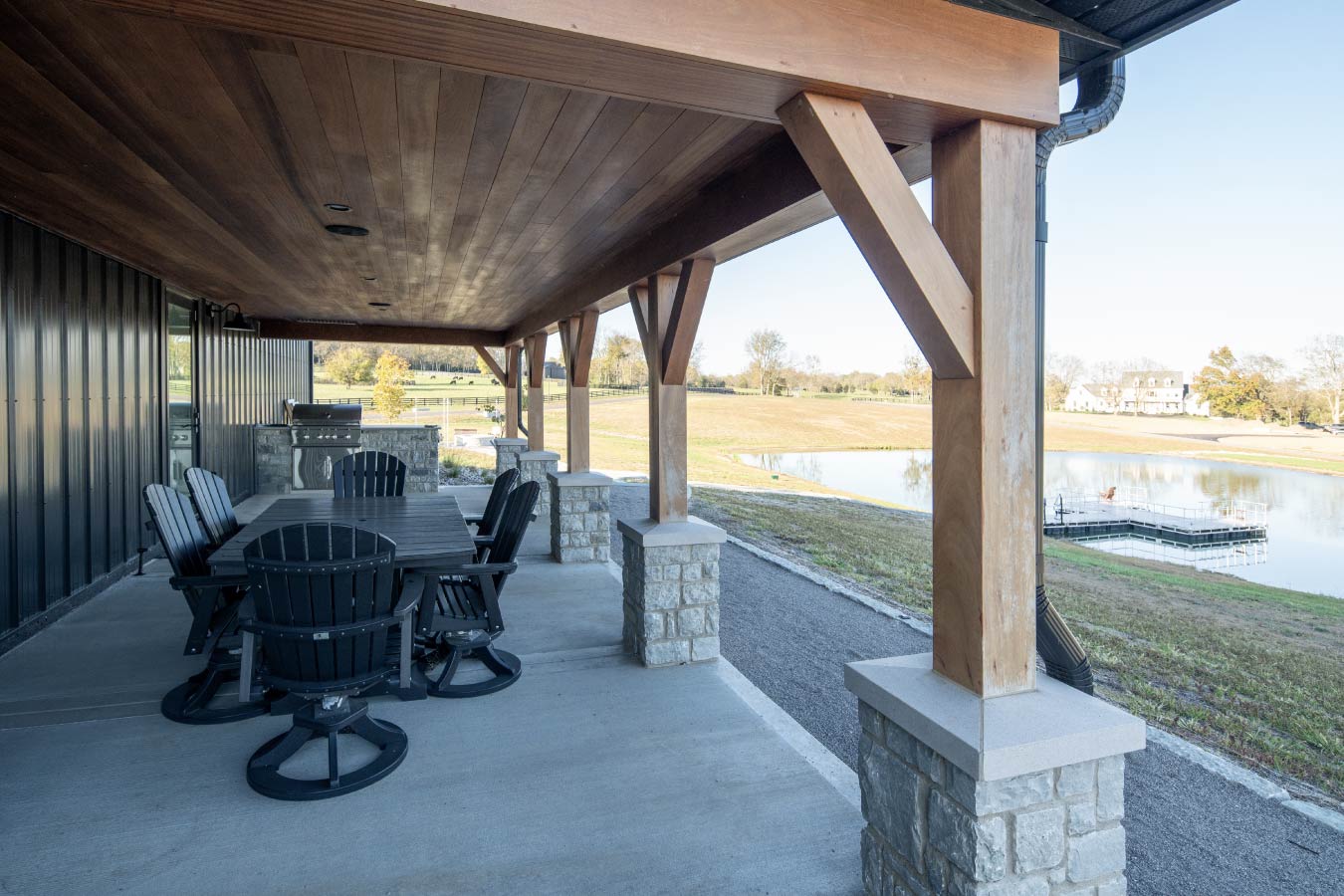 Covered patio at the Keene Ranch event barn with timber posts, outdoor dining table, stone columns, and pond views. Full View of Entertaining Porch
