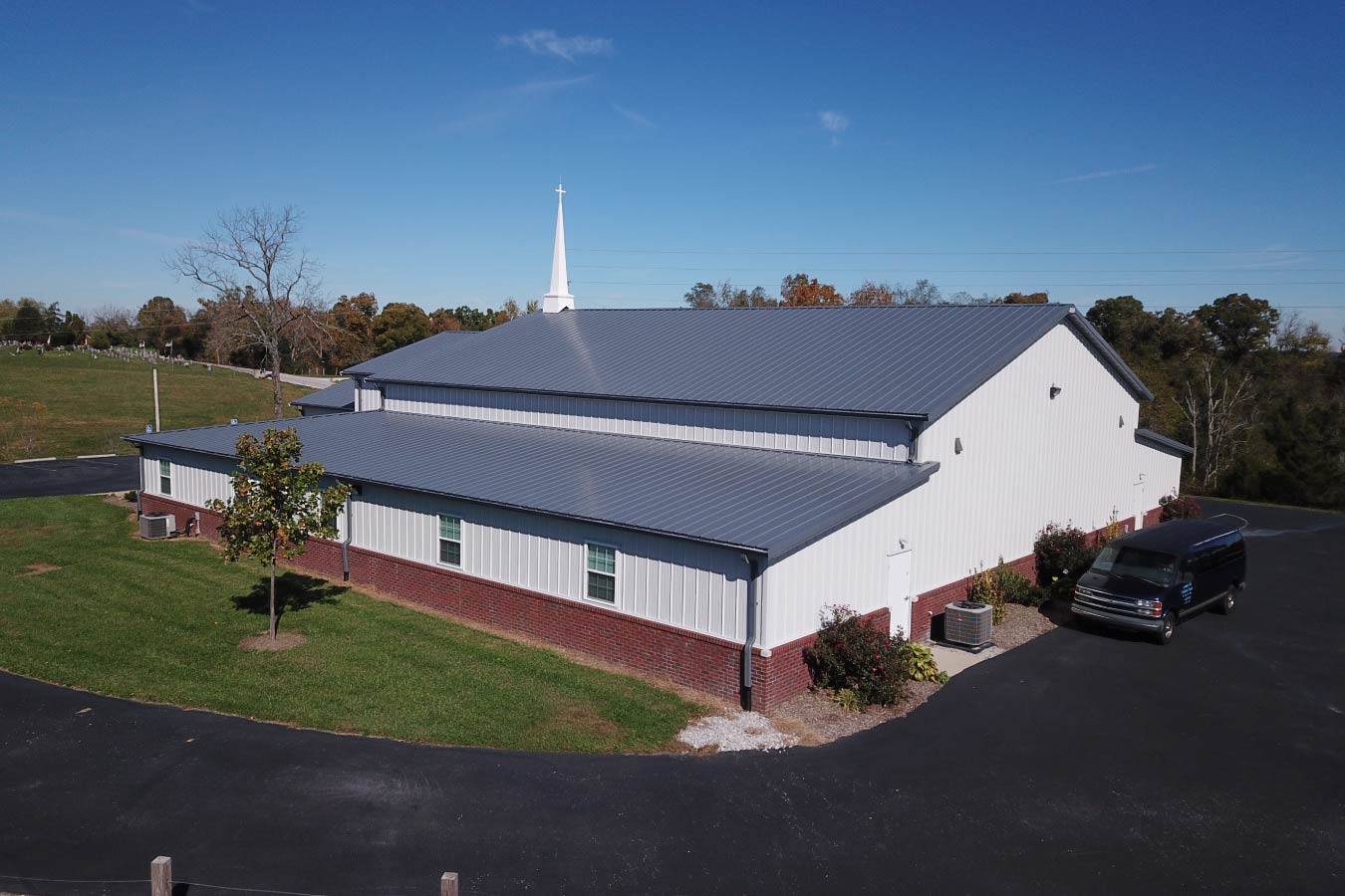 Side view of Pleasant Hill Christian Church with white steel siding, red brick wainscot, gray metal roof, and steeple. Aerial View of Back of Building