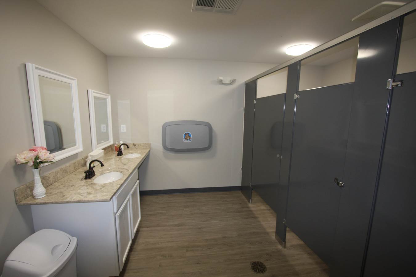 Modern commercial restroom interior with granite double vanity, black faucets, gray stall partitions, and wood-look flooring Modern Bathroom