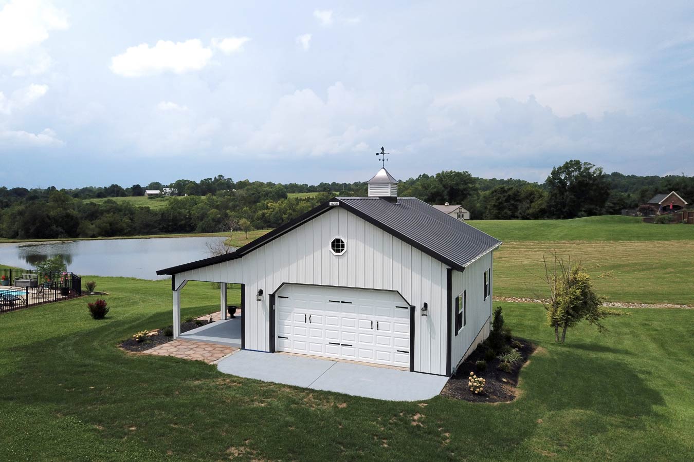 41-3783 pole shed with porch, white steel siding, black trim, garage door, and cupola near pond