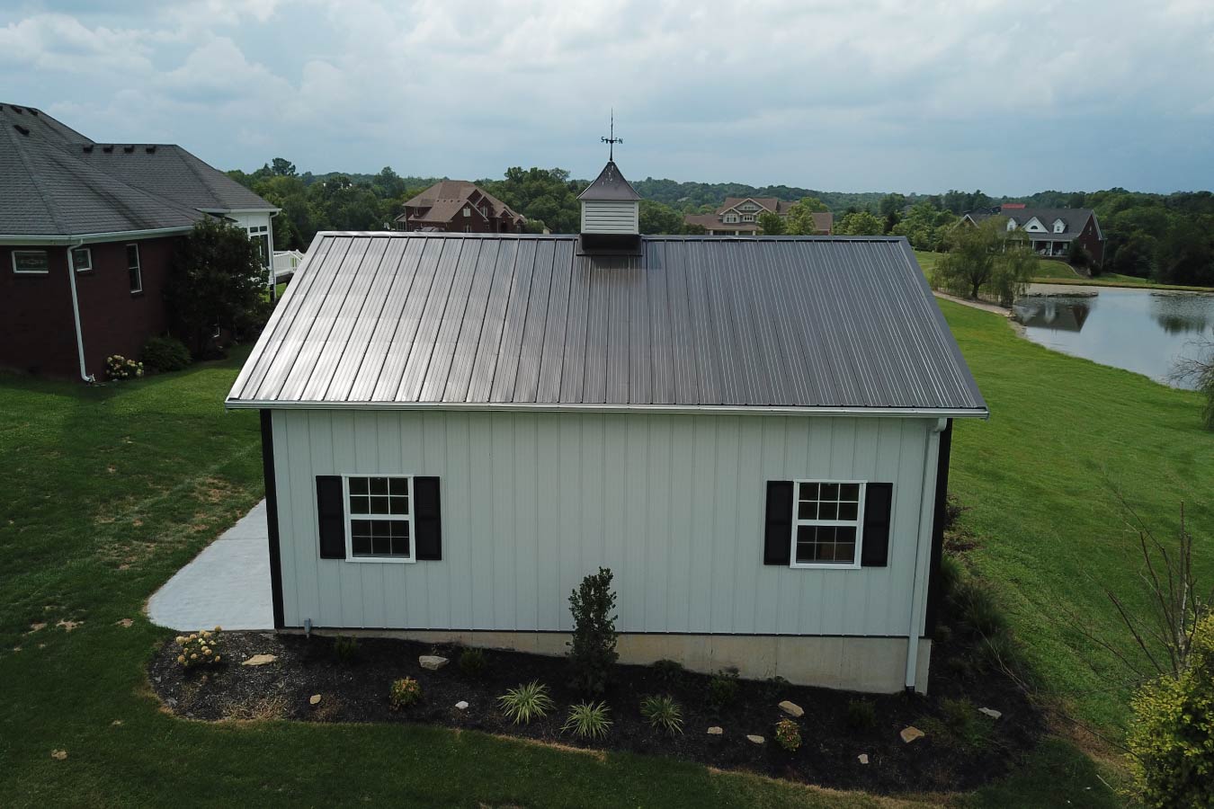 This pole shed combines functionality and curb appeal with a clean, modern design made possible through post frame construction. The building features white steel siding with black trim, creating a sharp contrast that stands out against the natural surroundings. A key feature of this pole shed is the covered porch, offering a comfortable outdoor space for relaxing or transitioning into the building. The overhead garage door provides easy access for equipment, tools, or vehicles, making the structure highly versatile. The addition of a cupola enhances ventilation while adding a classic architectural touch. The surrounding landscape, including the nearby pond and open green space, highlights how this pole shed integrates seamlessly into a rural property. Built with post frame construction, this pole shed delivers durability, low maintenance, and an open interior layout—making it ideal for storage, a workshop, or a multi-purpose outbuilding.