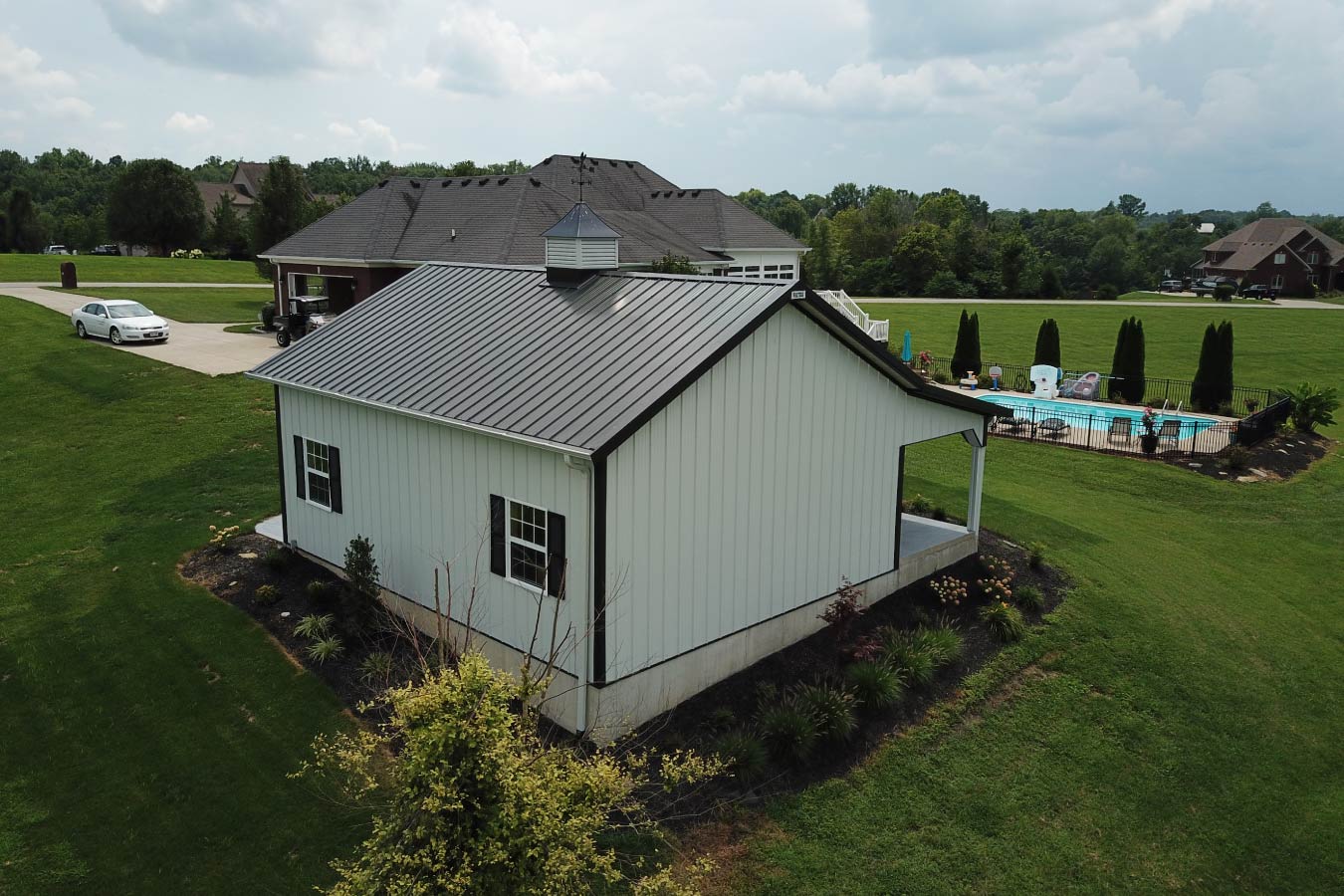 This pole shed showcases a clean and efficient design built using post frame construction, offering both durability and visual appeal. The white steel siding paired with black shutters creates a crisp, modern look that complements a variety of property styles. The metal roof provides long-lasting protection and low maintenance, while the centrally placed cupola enhances ventilation and adds a traditional architectural element. Two sidewall windows bring natural light into the interior, improving usability for storage or workspace applications. Surrounded by simple landscaping and open green space, this pole shed fits seamlessly into its environment while maintaining a strong visual presence. With the advantages of post frame construction, this building delivers a wide-open interior layout, making it ideal for storage, a workshop, or a versatile multi-purpose space.