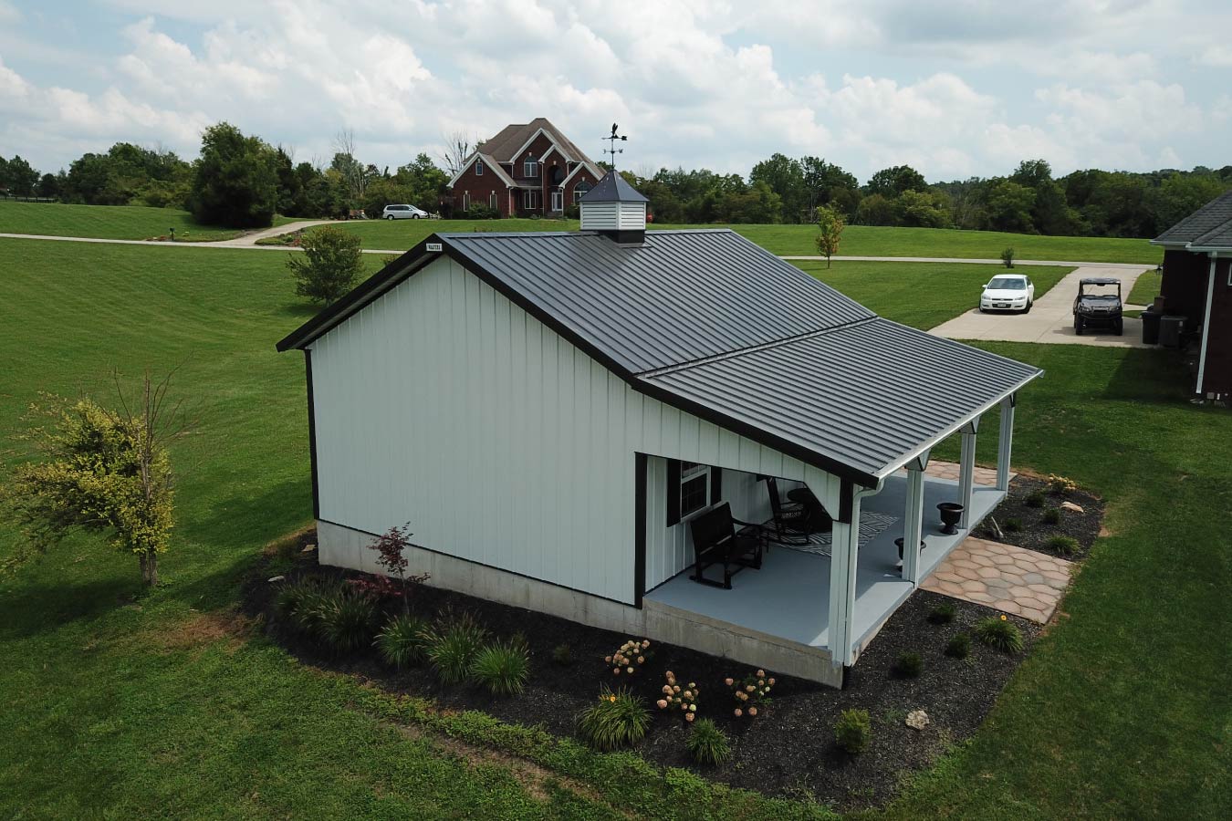 This pole shed highlights a clean, modern design paired with the durability of post frame construction. The white steel siding combined with black trim creates a sharp, high-contrast appearance that stands out while complementing the surrounding landscape. A key feature of this pole shed is the covered porch, providing a functional outdoor space for relaxing or transitioning into the building. The metal roof ensures long-term durability and low maintenance, while the centrally placed cupola enhances ventilation and adds a classic barn-style detail. Two sidewall windows allow natural light into the interior, improving usability for storage, workshop, or hobby applications. The surrounding landscaping and nearby amenities highlight how this pole shed integrates seamlessly into a residential setting. Built with post frame construction, this pole shed offers a wide-open interior layout, flexibility, and long-lasting performance—making it ideal for a variety of uses.