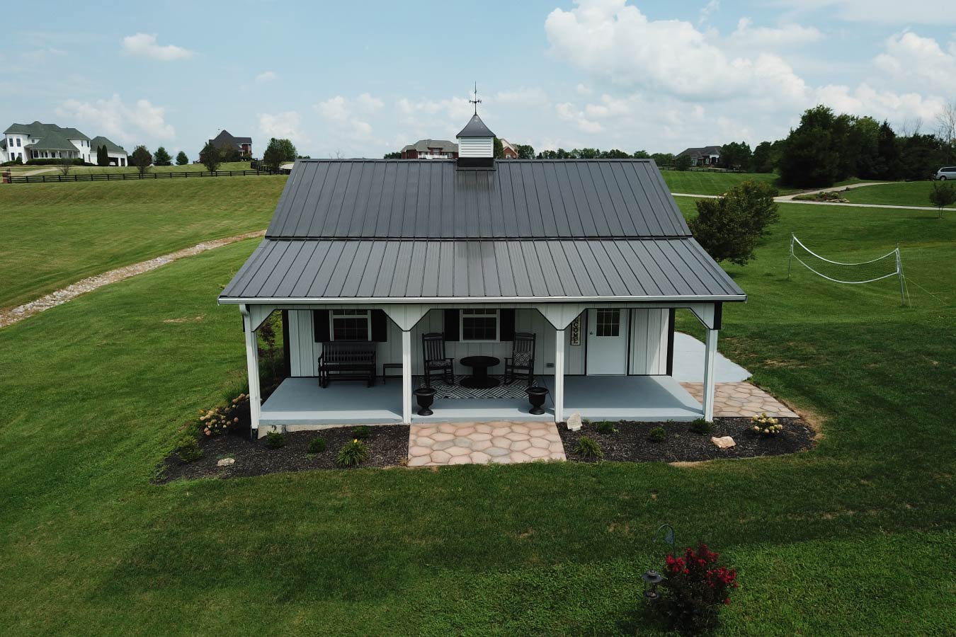 This pole shed combines modern design with the efficiency of post frame construction, making it a perfect fit for residential properties. The building features white steel siding accented with black trim, creating a clean and contemporary aesthetic. A prominent feature of this pole shed is the covered porch, supported by square posts and enhanced by decorative brackets. This space provides a functional outdoor seating area, ideal for relaxing while adding architectural interest to the structure. The gray metal roof delivers long-term durability and low maintenance, while the cupola with a weathervane adds both ventilation and a classic barn-style detail. The overhead garage door allows easy access for vehicles, equipment, or storage needs. Surrounded by professional landscaping and set on a concrete slab foundation, this pole shed demonstrates how a practical structure can also elevate the look of a property. Its versatile layout makes it ideal for use as a garage, workshop, or hobby space.