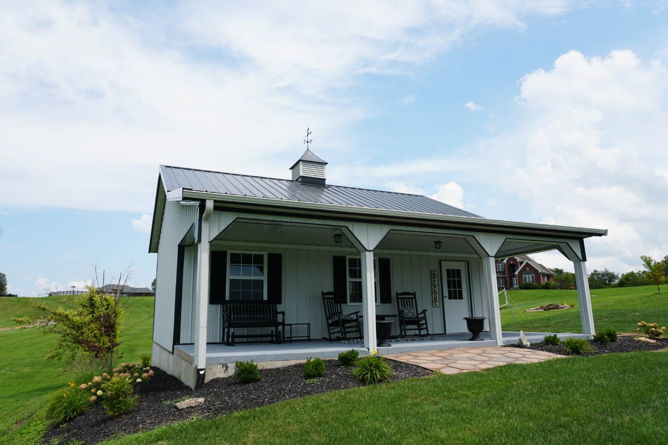 This pole shed features a symmetrical front design that blends functionality with clean, modern aesthetics. Built using post frame construction, the structure showcases white steel siding with bold black trim, creating a sharp and timeless exterior. The standout feature is the full-length covered porch, supported by evenly spaced posts and enhanced with decorative brackets. This outdoor space provides a comfortable area for seating and relaxation while adding depth and character to the building’s façade. The gray metal roof ensures durability and low maintenance, while the centrally positioned cupola with weathervane adds ventilation and a traditional barn-style accent. A concrete slab foundation and professionally landscaped surroundings further elevate the appearance and usability of this pole shed. Designed for versatility, this pole shed can function as a garage, workshop, or hobby space, offering both practical performance and strong curb appeal through efficient post frame construction.