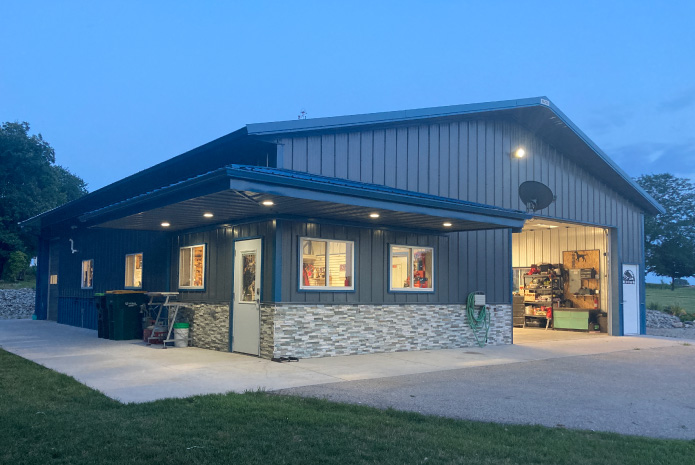 Lake House Garage with Covered Porch and Stone Wainscot at Dusk | Walters Buildings Lake house garage at dusk with steel siding, stone wainscot, covered porch, and open overhead door