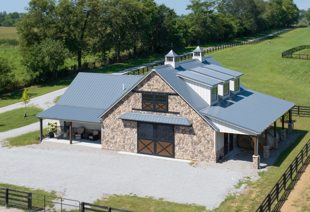 Post-frame horse barn with stone exterior, steel roof, cupolas, and covered porches in rural setting