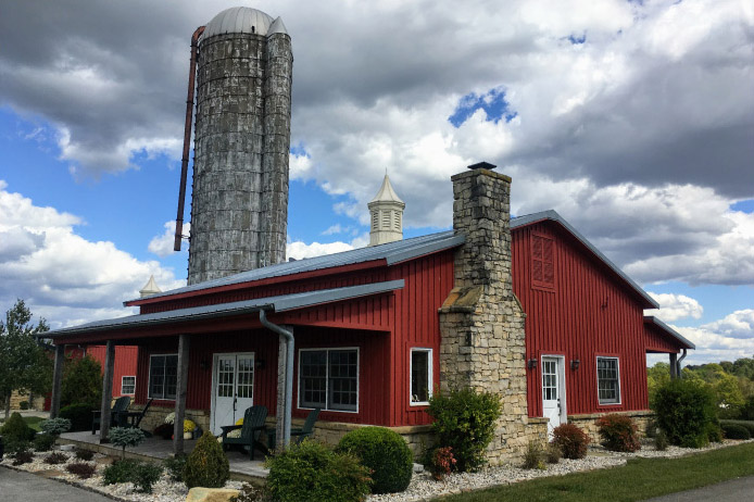 Pole barn house with red steel siding, metal roof, covered porch, stone chimney, and farm silo