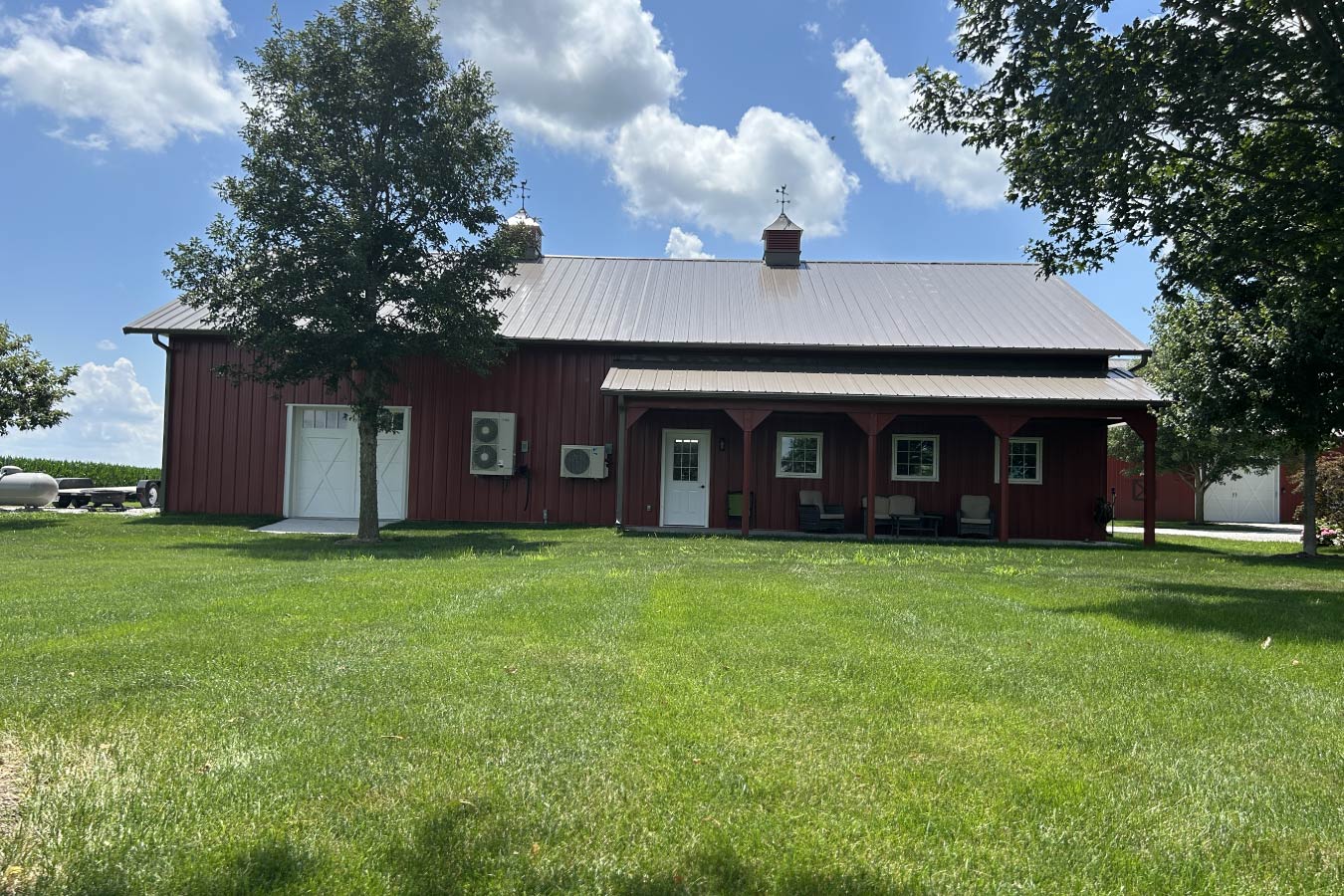 Residential workshop with red steel siding, steel roof, covered porch, and single overhead door in landscaped yard Covered 8′×32′ rear porch designed for relaxation