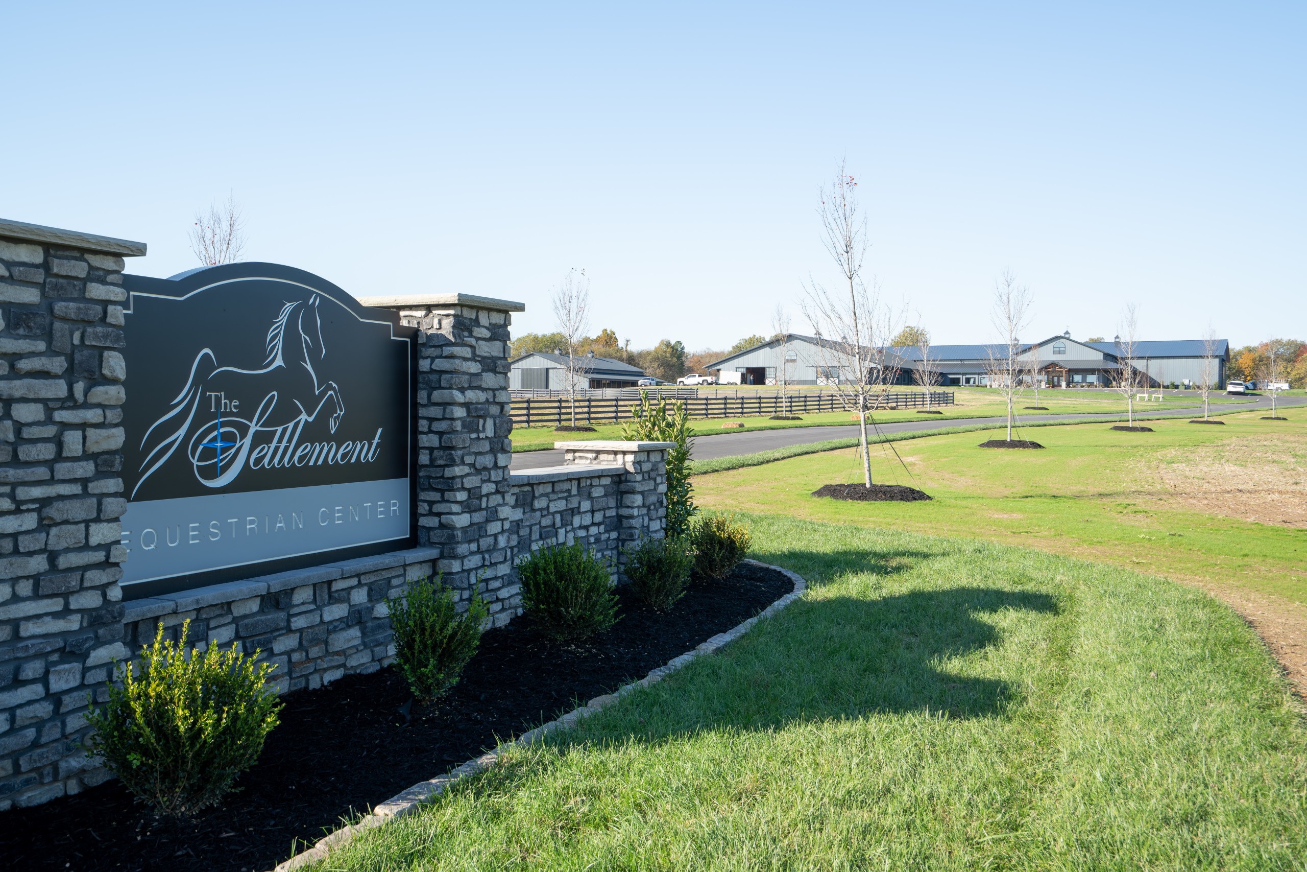 Stone entrance sign at custom equestrian center with horse barn and indoor riding arena in the background