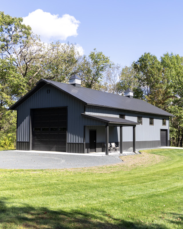 Custom 30×64 post frame garage with black metal siding and gable roof, surrounded by trees on a landscaped lot Custom 30x64 post frame garage with black metal siding and gable roof, surrounded by trees on a landscaped lot