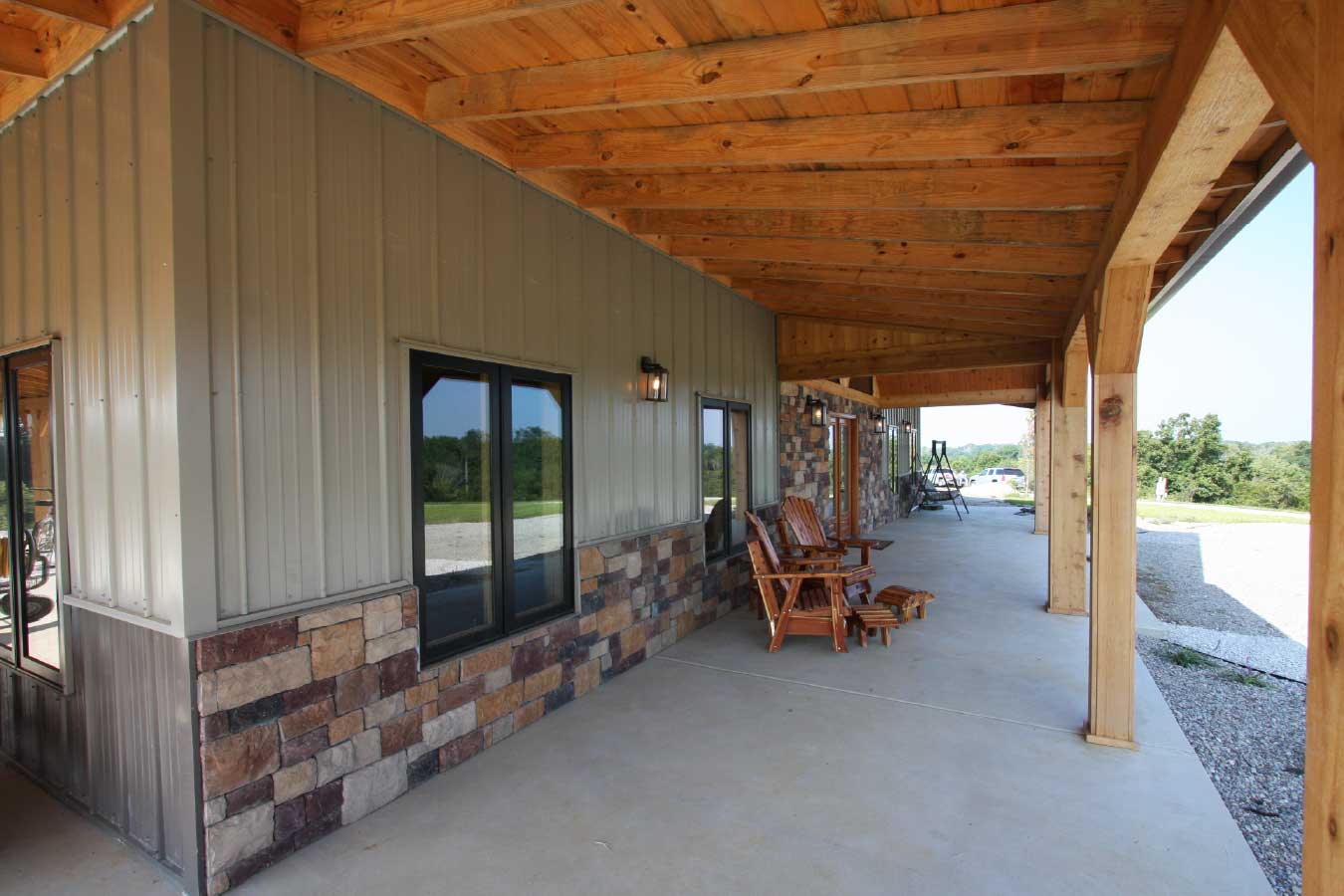 Close-up view under the wraparound porch of a luxury timber frame home, featuring steel siding, stone wainscot, and rustic wood ceiling details.