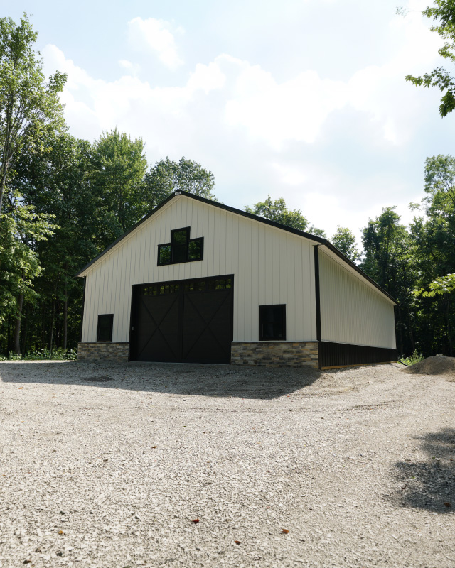 White post-frame storage building with black trim, cupola, and metal roof on gravel lot in wooded setting White post-frame storage building with black trim, cupola, and metal roof on gravel lot in wooded setting