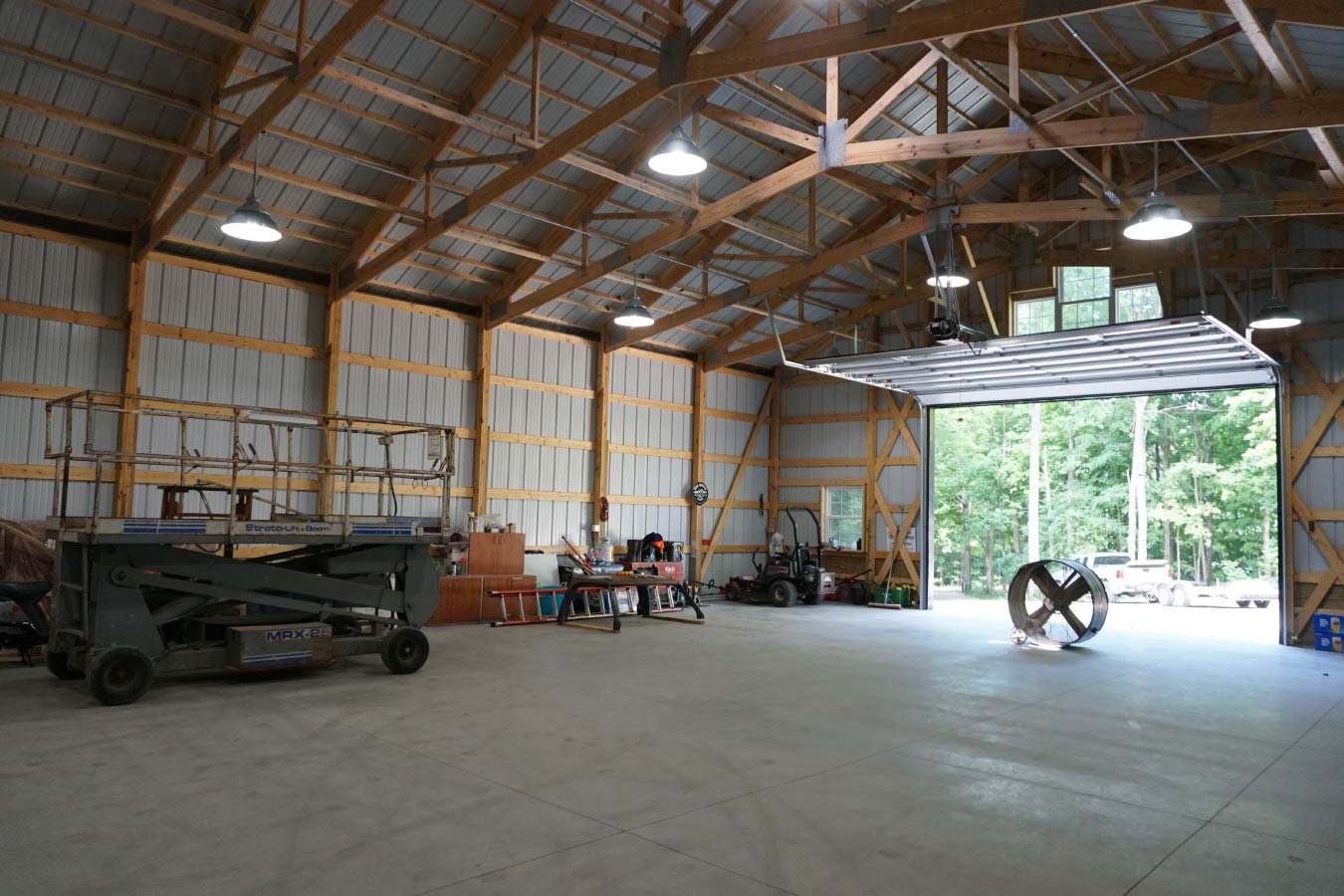 Post-frame building interior with exposed wood trusses, overhead lighting, and large open workshop space