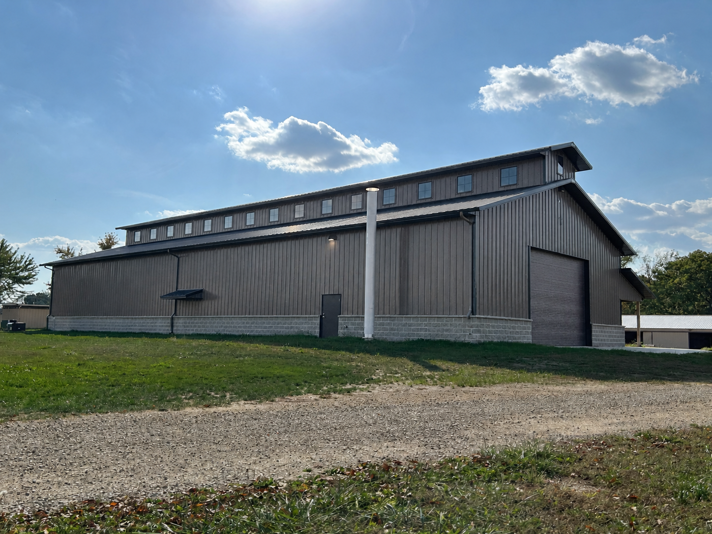 Side elevation of the Baer Powerhouse at Rock River Thresheree showing the monitor-style roofline, bronze siding, stone wainscot, exterior exhaust stack, and end-wall overhead door.
