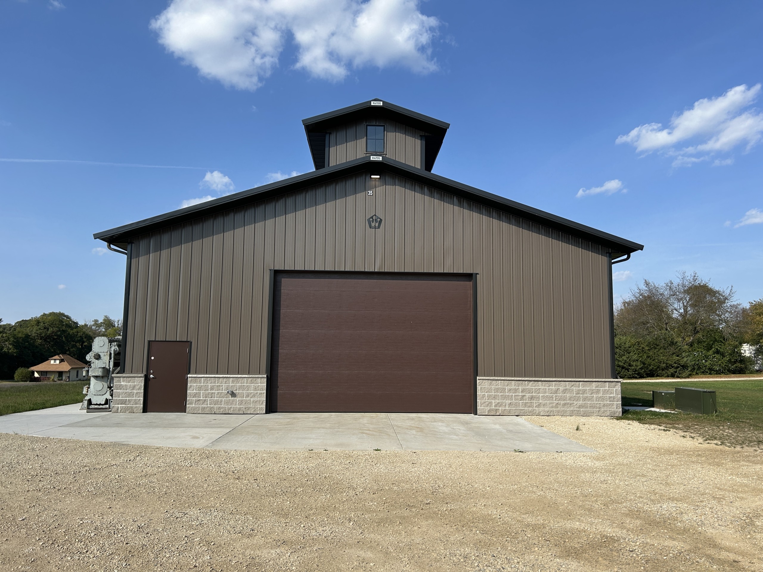 End view of the Baer Powerhouse at Rock River Thresheree with a 20′ × 14′ overhead door, bronze siding, black roof, stone wainscot, and monitor-style roofline.