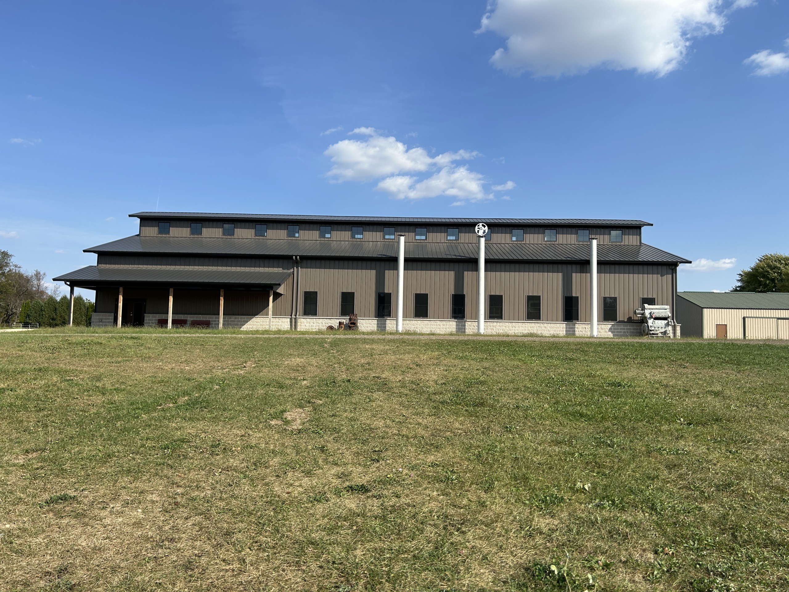 Side view of the Baer Powerhouse at Rock River Thresheree with exterior exhaust stacks, covered porch, monitor-style roofline, bronze siding, and stone wainscot.