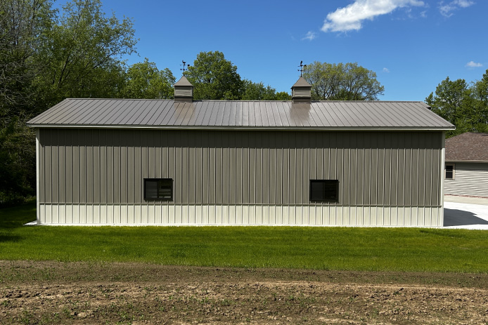 L shaped pole barn with brown steel siding, white wainscot, black windows, and decorative cupolas
