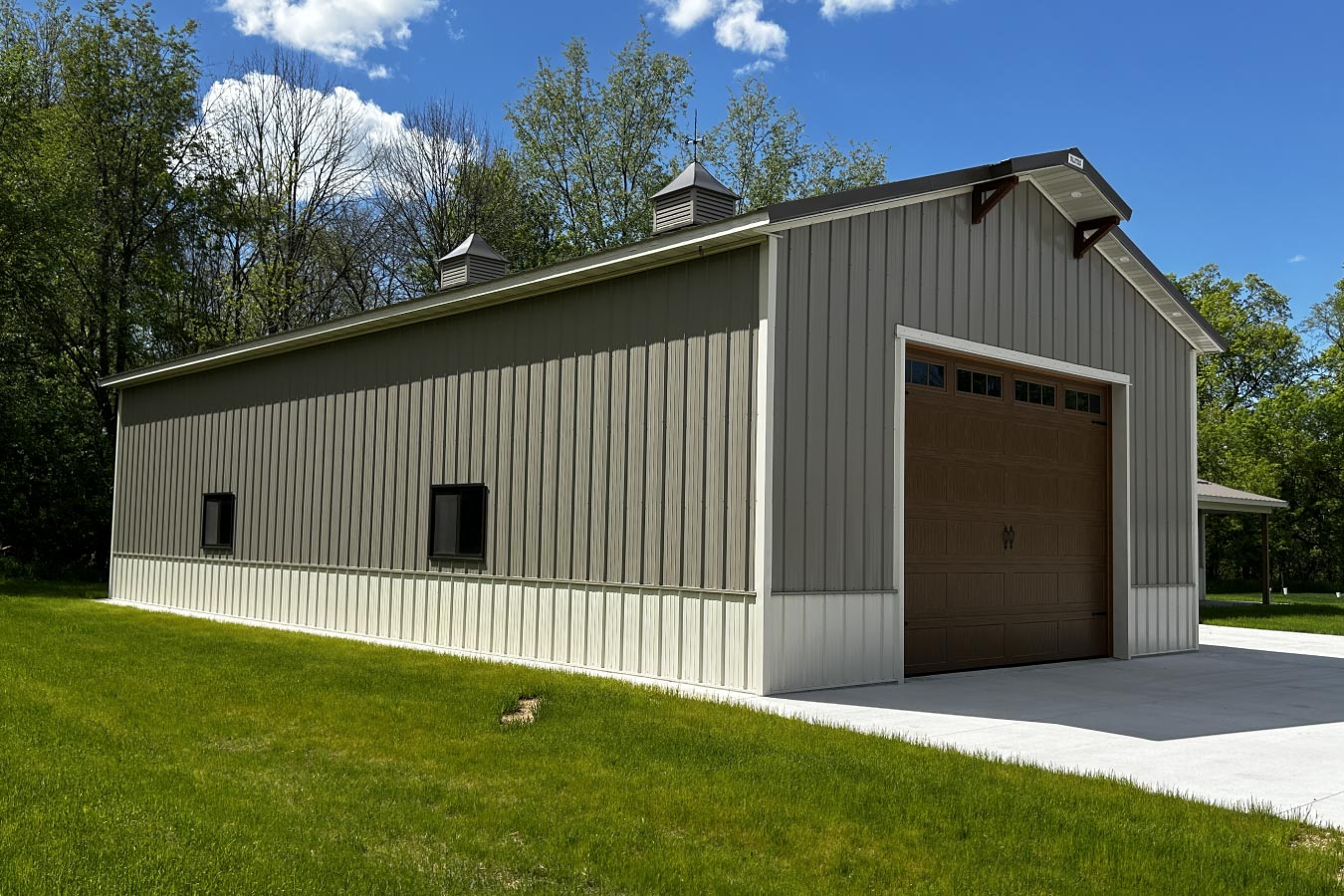 L shaped pole barn garage with gray steel siding, white wainscot, cupolas, and oversized brown garage door