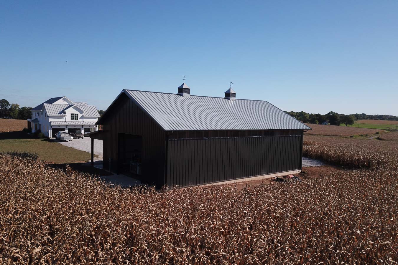 Rear view of 40x64 pole barn on acreage with black steel siding, cupolas, and custom home in the background