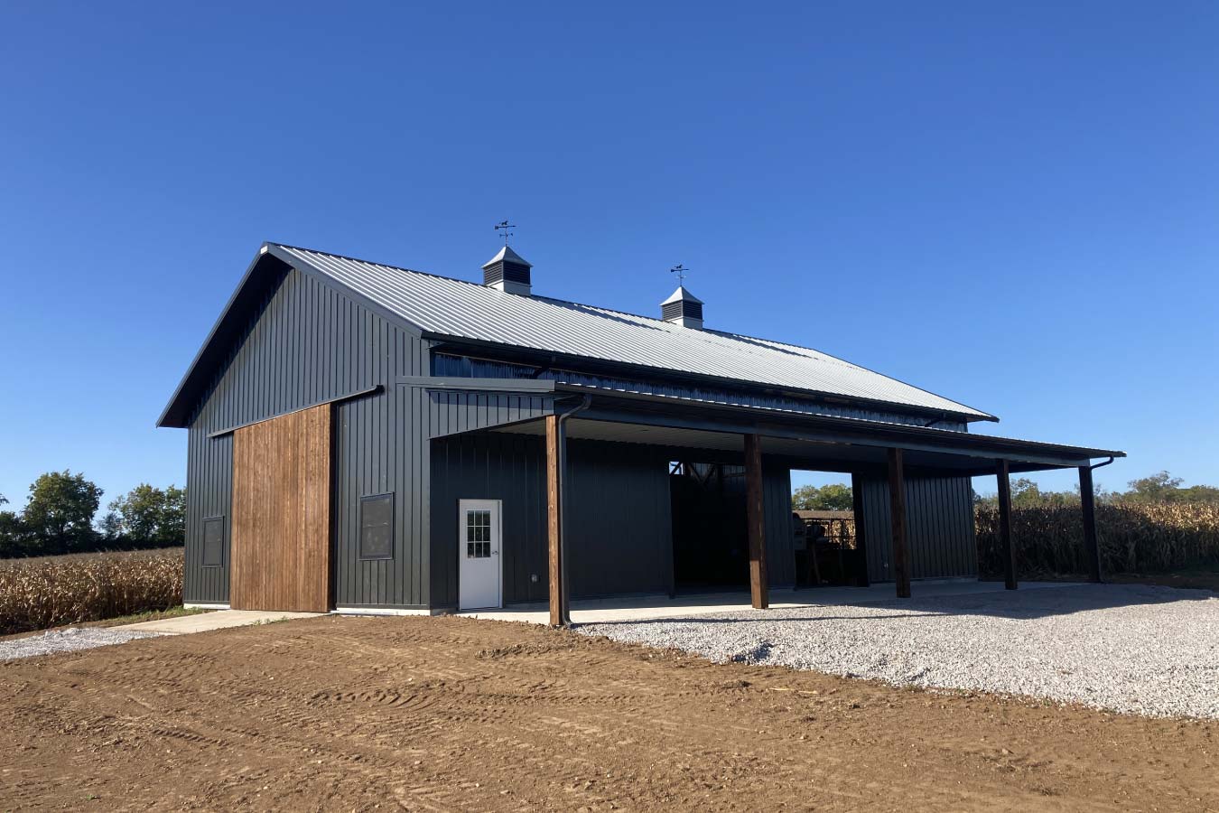 Angled view of post-frame pole barn with lean-to addition, wood posts, and black metal siding in rural setting