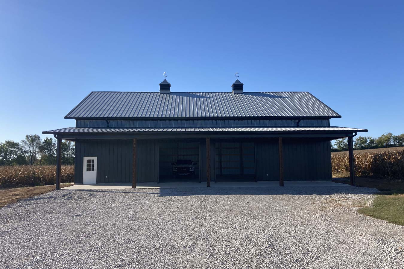 Front view of pole barn with full-width lean-to, gravel drive, and open bays for equipment and vehicle storage