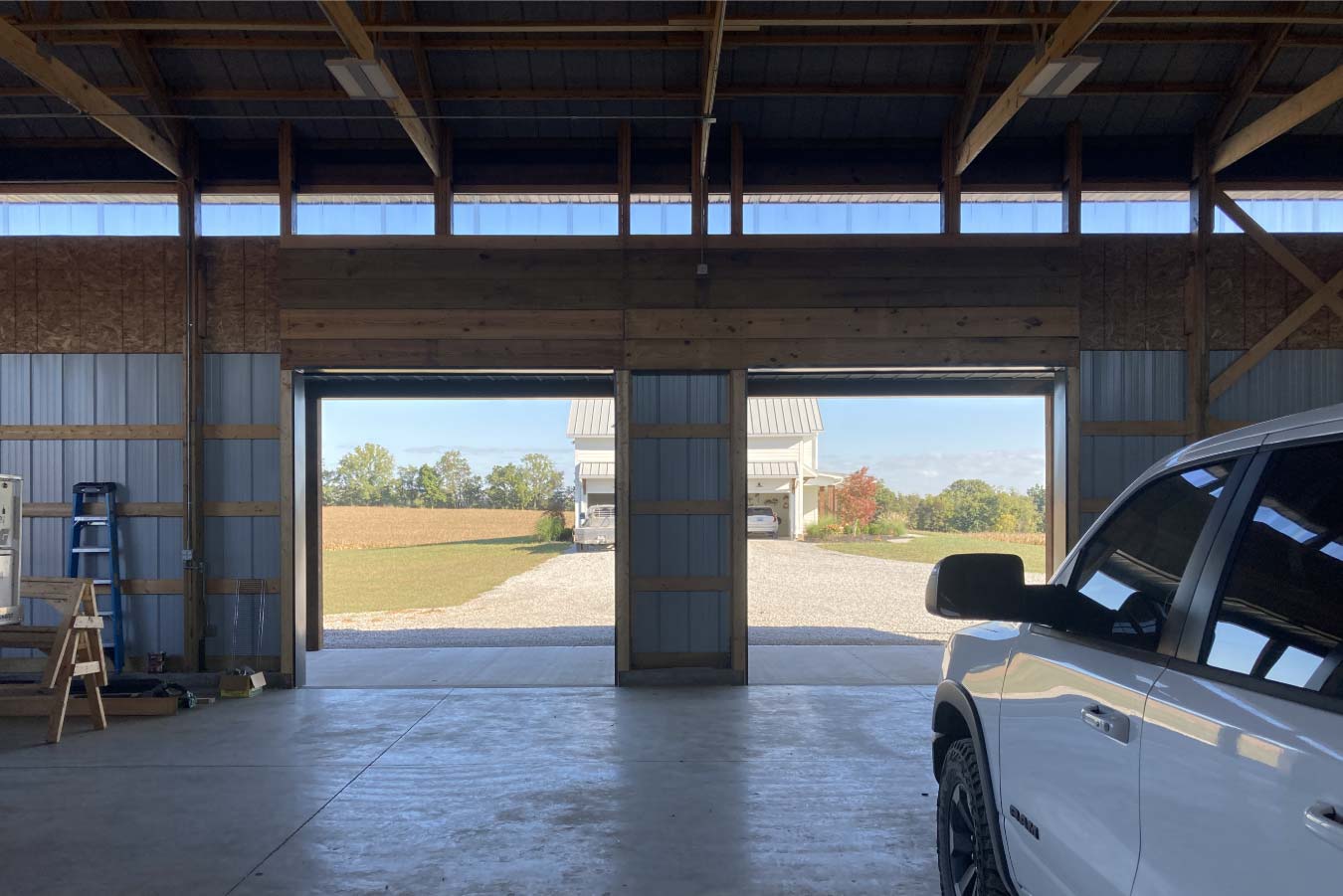 Interior view from inside pole barn looking out through large openings toward gravel drive and nearby home